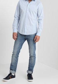 Světle modrá košile na knoflíky s dlouhými rukávy, zkombinovaná se světlými modrými džíny a černými teniskami s bílou podešví a hnědými akcenty.