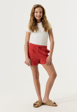 Lachend meisje met lang krullend haar, gekleed in een wit T-shirt met korte mouwen, rode shorts met trekkoord en beige sandalen, staand voor een effen achtergrond.