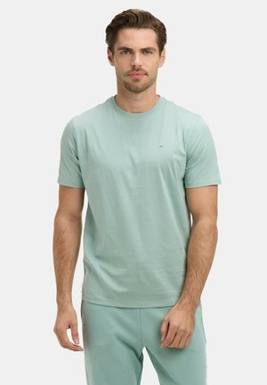 Homme portant un T-shirt à manches courtes vert clair et un pantalon assorti, debout devant un fond uni clair.
