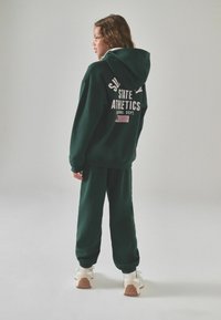 Felpa con cappuccio verde con la scritta "STATE ATHLETICS" sulla schiena, abbinata a pantaloni jogger coordinati. Caratterizzata da una vestibilità comoda e da una texture morbida.
