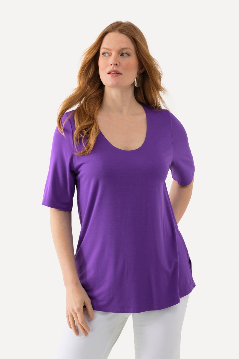 Femme aux longs cheveux roux portant un haut violet à manches courtes et un pantalon blanc, regardant légèrement vers sa droite sur un fond uni.