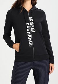 Felpa nera con zip realizzata in tessuto morbido, dotata di tasca a marsupio frontale e logo bianco "ARMANI EXCHANGE" che corre in verticale.