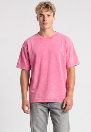 Giovane uomo con capelli biondi ricci che indossa una t-shirt rosa a maniche corte e jeans grigi, in piedi davanti a uno sfondo chiaro e semplice.