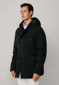 Muškarac u crnom zimskoj kaputu s kapuljačom preko prugaste košulje, stoji naslonjen na jednostavnu svijetlu sivu pozadinu.