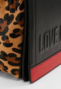 Gros plan sur le coin d'un sac en fourrure léopard, panneau en cuir noir embossé avec "LOVE J", et une bande en cuir rouge à la base.