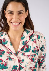 Donna sorridente con capelli scuri che indossa una maglietta da pigiama bianca con motivi floreali verdi e rosa e rifiniture bordeaux.