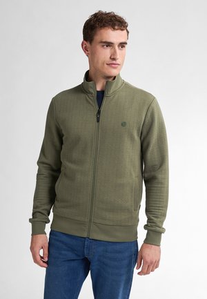 Olive green zip-up jacket with a textured pattern. Features a high collar, long sleeves, and ribbed cuffs. Worn over a dark shirt.