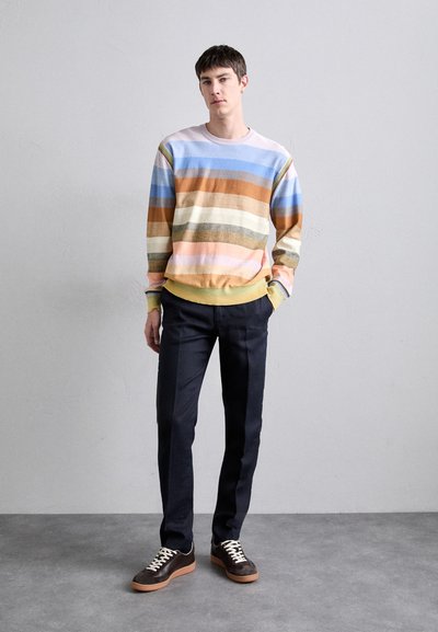 Pulôver listrado multicolorido em tons pastel de azul, rosa, castanho e creme, com uma bainha amarela; combinado com calças escuras de corte clássico e ténis castanhos.