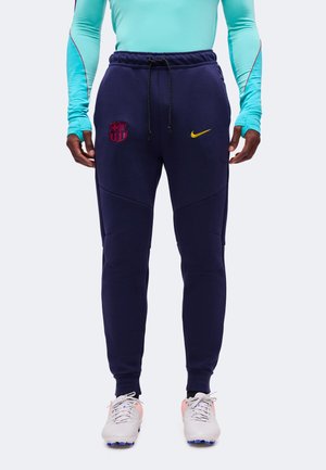 Mann trägt marineblaue Jogginghosen mit FC Barcelona und gelben Nike-Logos, kombiniert mit weißen Fußballschuhen und einem hellblauen Langarmshirt.