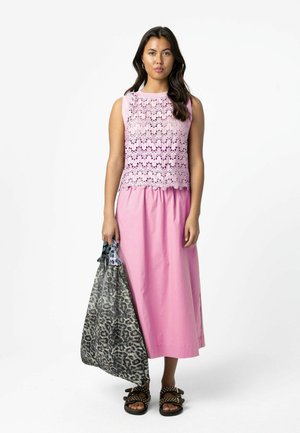 Žena v svetloružovom čipkovanom tielku bez rukávov a zodpovedajúcej ružovej sukni, drží veľkú čierno-bielo vzorovanú tašku, na nohách má čierne sandále.