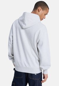 Sudadera de algodón gris con capucha ajustable, puños y cintura acanalados. Textura suave, corte relajado, con una etiqueta de logo cerca de la cintura.