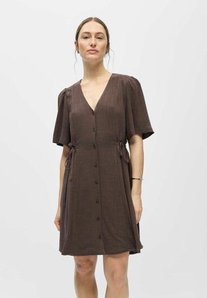 Vrouw draagt een bruine jurk met korte mouwen en knopen aan de voorkant, met zijstrikjes, staand tegen een effen lichte achtergrond.