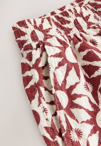 Gros plan d’un tissu de ceinture élastique avec des motifs de soleil rouges sur fond blanc, présentant un matériau texturé, froncé et une couture latérale visible.