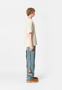 Camiseta beige de corte oversized, jeans de mezclilla azul con parches laterales y perro bordado, zapatos color tostado. Diseño sencillo, ajuste relajado, textura suave.