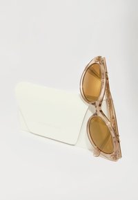 Ochelari de soare transparenți cu lentile maro, având un design îndrăzneț, rotund, și accente intricate; acompaniați de o carcasă moale albă.