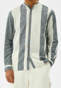 Overhemd met verticale blauw-witte strepen, linnen structuur, bandkraag en knopen in houtkleur, gecombineerd met beige broek.