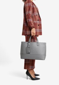 Bolso tote de cuero sintético gris con asas dobles, una etiqueta colgante y costuras sutiles, combinado con un traje de cuadros y tacones negros.