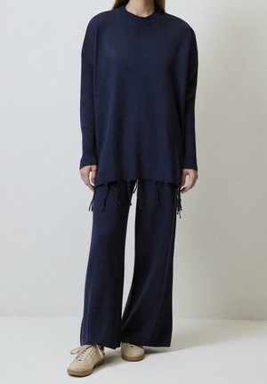 Persoon draagt een marineblauwe oversized gebreide trui met franjes aan de zoom en bijpassende wijde broek, gecombineerd met beige sneakers.