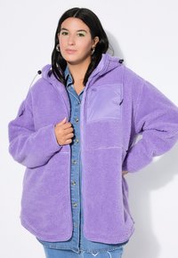 Lila Fleecejacke mit Kapuze, ausgestattet mit einer Reißverschlusstasche auf der Brust und weichem Material. Trägt man über einem Jeanshemd mit Knopfverschluss.