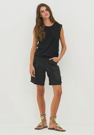 Femme aux longs cheveux bruns portant un haut noir sans manches, un short cargo noir avec des poches, et des sandales cloutées dorées, debout devant un fond blanc.