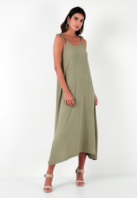 Olijfgroene, mouwloze maxi-jurk van een textuurstof, met verstelbare schouderbandjes en een high-low zoom. Gecombineerd met beige sandaaltjes.