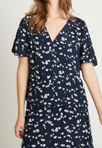 Blusa floreale blu navy con maniche corte a sbuffo; presenta un scollo a V, chiusura con bottoni e piccoli motivi floreali bianchi, rosa e blu.