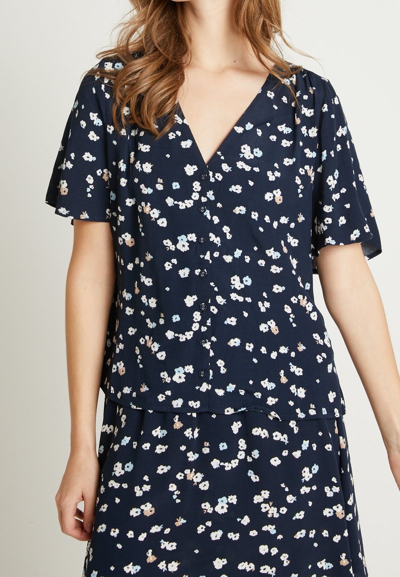 Blusa floreale blu navy con maniche corte a sbuffo; presenta un scollo a V, chiusura con bottoni e piccoli motivi floreali bianchi, rosa e blu.