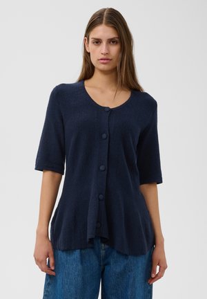 Jeune femme portant un cardigan bleu marine à manches courtes boutonné et un jean bleu, debout devant un fond clair uni.
