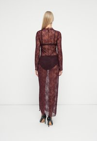 Sieviete ar gariem blondiem matiem, tērpta caurspīdīgā bordo krāsas mežģīņu kleitā virs atbilstošām apakšveļas komplekta un melniem augstpapēžu apaviem, pagriezusies prom.