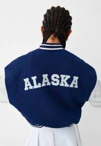 Blå varsityjacka med vita ärmar, med "ALASKA" broderat på ryggen i ljusblått, ribbat krage och fåll. Strukturerad tyg.