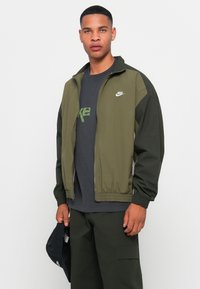 Olivno zelena zapinjalna jakna z elastičnimi manšetami in pasom, ki ima bel Nike logo. Oblačena je čez temno srajco in v kombinaciji z zelenimi hlačami.