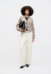 Šviesiai rudos spalvos megztas kardiganas su mygtukais, derinamas su kremine spalvos plačiomis kelnėmis ir juodais smailiais aukštakulniais, laikantis juodą delninę.