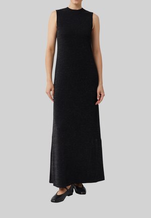 Czarna sukienka maxi bez rękawów z delikatnym połyskiem, noszona z czarnymi płaskimi butami z paskami skrzyżowanymi i wycięciami.