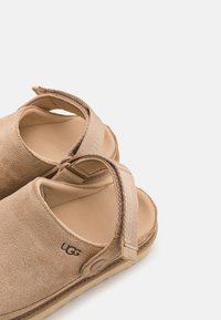 Smeđe suede japanke bez remena s prilagodljivim trakama, podstavljenim uloškom i UGG logom. Teksturirana površina i fleksibilna gumena potplat.