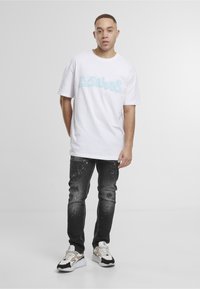 Jeune homme debout, portant un t-shirt blanc graphique, un jean noir déchiré avec des éclaboussures de peinture et des baskets blanches et beiges.