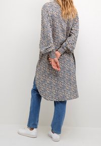Vestido camisero de manga larga en estampado floral beige y azul, con un corte relajado, dobladillo redondeado y puños fruncidos. Combinado con jeans azules y zapatillas blancas.