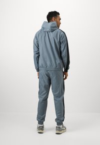 Lettet gråt tracksuit med hætte, elastiske manchetter og sorte sidepaneler. Har en afslappet pasform og synlige stikninger.