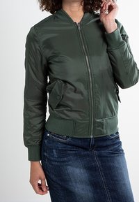 Zielona bomberka wykonana z błyszczącego, gładkiego materiału z prążkowanym kołnierzem i mankietami; posiada zamek błyskawiczny i boczną kieszeń.