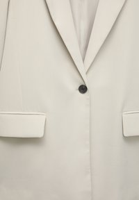 Beige Blazer aus glattem Stoff mit einem einzelnen schwarzen Knopf, Reverskragen und zwei Vorderseiten Taschen mit umgeschlagenen Ärmeln.