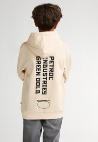 Sudadera color crema fabricada con una mezcla de algodón, con un gran texto negro "PETROL INDUSTRIES GREEN GOLD" en vertical en la parte posterior.