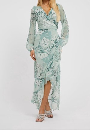 Femme portant une robe portefeuille fleurie vert clair avec des manches longues en voile et un ourlet asymétrique à volants, assortie à des talons ouverts scintillants.