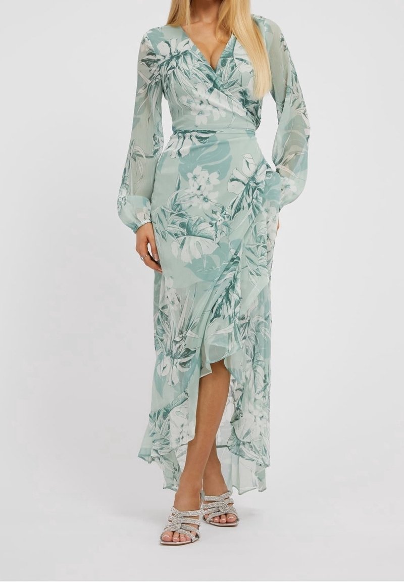 Femme portant une robe portefeuille fleurie vert clair avec des manches longues en voile et un ourlet asymétrique à volants, assortie à des talons ouverts scintillants.