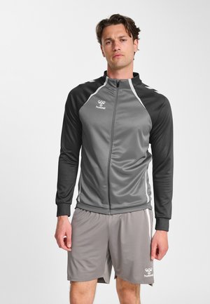 Homme debout portant une veste de sport zippée grise et noire et un short de sport gris assorti avec des logos de marque sur la poitrine et la jambe.