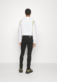 Bílá košile s dlouhým rukávem a výraznými vzorovanými šlemi, doplněná přiléhavými černými kalhotami a teniskami se zlatými detaily, pohled zezadu.