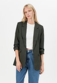 Šedý blazer se strukturovaným designem, klopovým límcem, dlouhými rukávy s ohrnutými manžetami a dvěma předními kapsami; nošený přes bílý top a modré džíny.