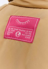 Ružová textilná nálepka s bielou vyšitou zavretou okom, ikonou Zeme a textom na béžovom odevnom texte.