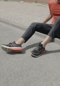 Laufschuhe: schwarzes Mesh-Obermaterial mit roten und grünen Akzenten, gepolsterte Sohle, flacher Schnürverschluss, auf einem grauen Gehweg neben einem Bordstein.
