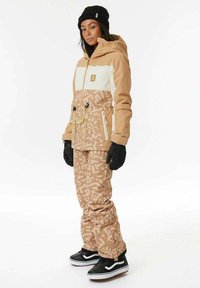 Giacca da sci beige e bianca con cappuccio, caratterizzata da una sezione inferiore a motivo, abbinata a pantaloni beige a motivo e guanti e scarpe neri.