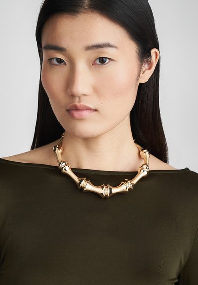 Zlatý náhrdelník se skládáním trubkového designu, který obsahuje zakřivené a ostré články, nošený přes tmavě zelený top bez ramínek.