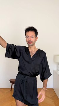 Črni satenast plašč s širokimi rokavi in pasom, ki se veže, z gladko teksturo in rahlim sijajem, nošen preko svetle barve kože.